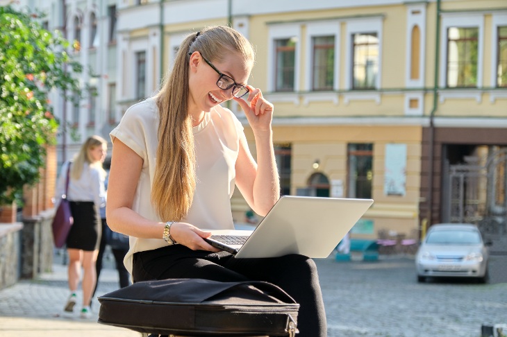 Devojka sedi napolju gleda u laptop dok jednom rukom pridržava naočare za kompjuter.